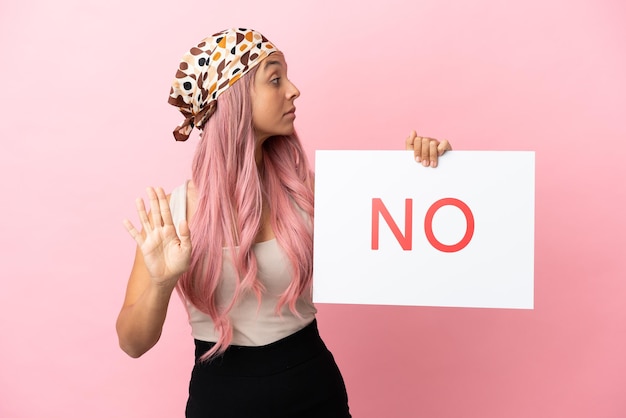 Young mixed race woman with pink hair isolated on pink background holding a placard with text NO and doing stop sign