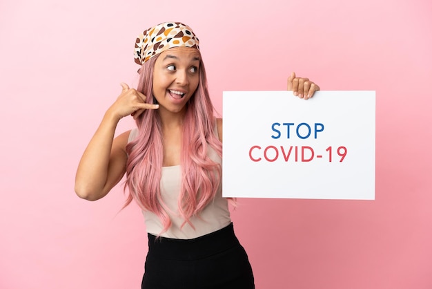 Young mixed race woman with pink hair isolated on pink background holding a placard with text Stop Covid 19 and doing phone gesture