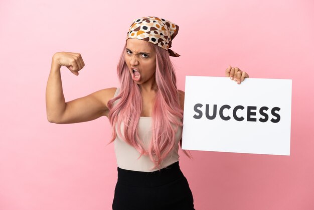 Young mixed race woman with pink hair isolated on pink background holding a placard with text SUCCESS and doing strong gesture