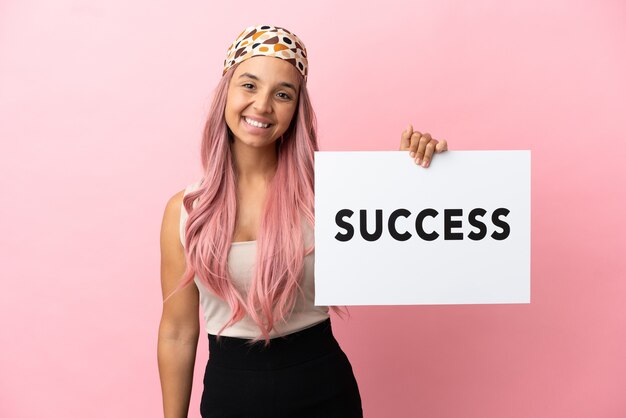 분홍색 배경에 격리된 분홍색 머리를 한 젊은 혼혈 여성이 행복한 표정으로 SUCCESS라는 문구가 적힌 플래카드를 들고 있습니다.