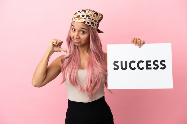 Foto giovane donna di razza mista con capelli rosa isolata su sfondo rosa con in mano un cartello con testo successo con gesto orgoglioso