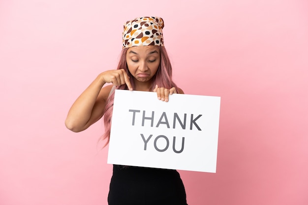 Young mixed race woman with pink hair isolated on pink background holding a placard with text THANK YOU and  pointing it