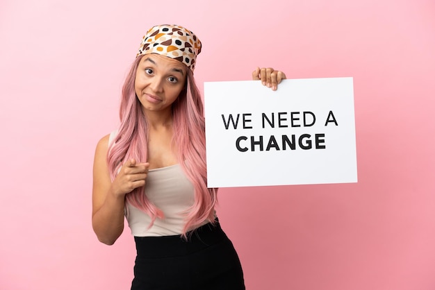 Молодая женщина смешанной расы с розовыми волосами на розовом фоне держит плакат с текстом «Нам нужны перемены» и указывает вперед