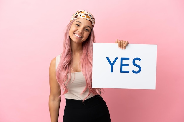 Young mixed race woman with pink hair isolated on pink background holding a placard with text YES with happy expression