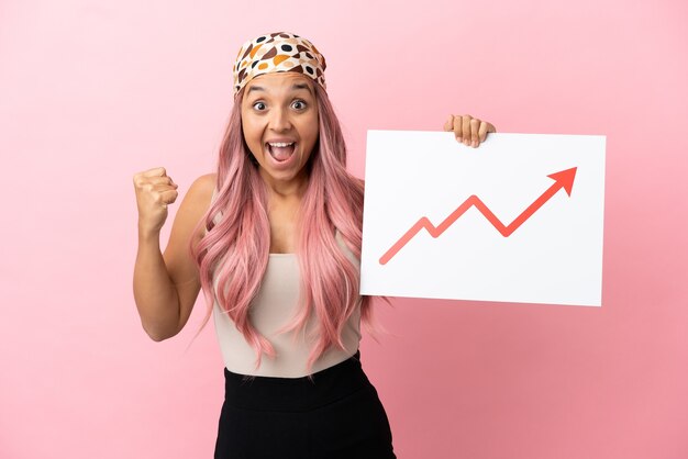 young mixed race woman with pink hair isolated on pink background holding a sign with a growing statistics arrow symbol and celebrating a victory