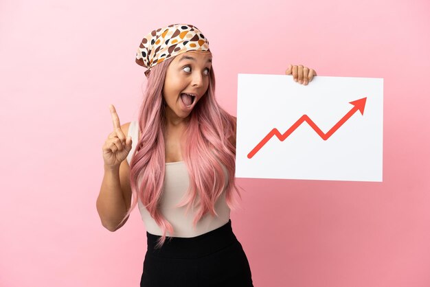 young mixed race woman with pink hair isolated on pink background holding a sign with a growing statistics arrow symbol and thinking