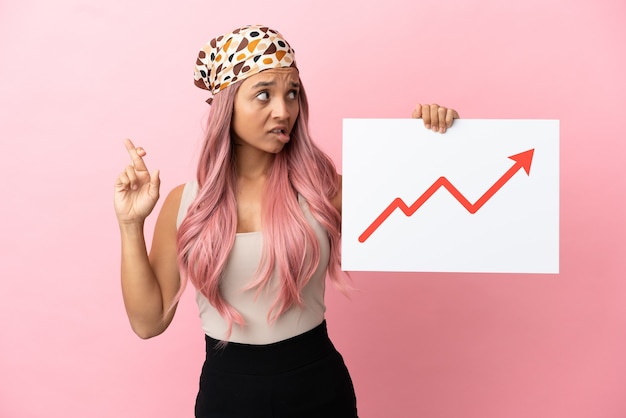 Young mixed race woman with pink hair isolated on pink background holding a sign with a growing statistics arrow symbol with fingers crossing