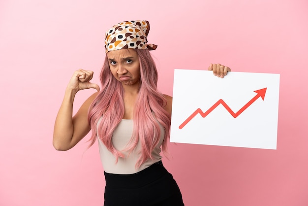 Foto giovane donna di razza mista con capelli rosa isolata su sfondo rosa con in mano un cartello con un simbolo di freccia statistica in crescita con gesto orgoglioso