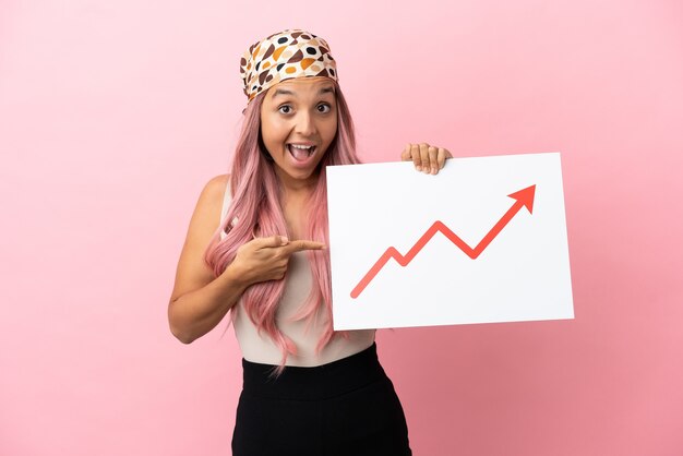 young mixed race woman with pink hair isolated on pink background holding a sign with a growing statistics arrow symbol with surprised expression