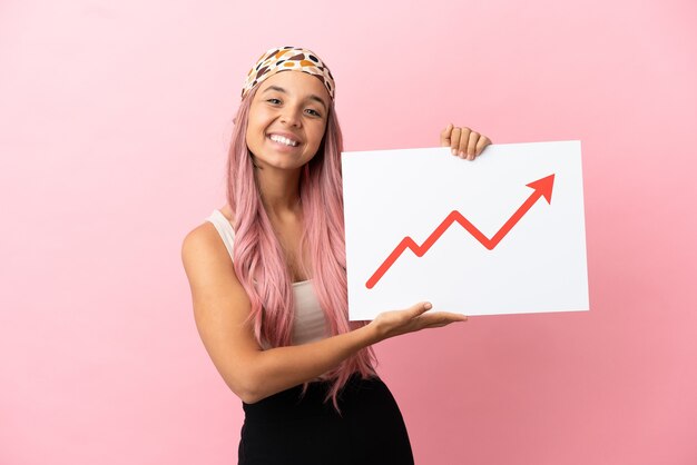 Giovane donna di razza mista con capelli rosa isolata su sfondo rosa con in mano un cartello con un simbolo di freccia statistica in crescita