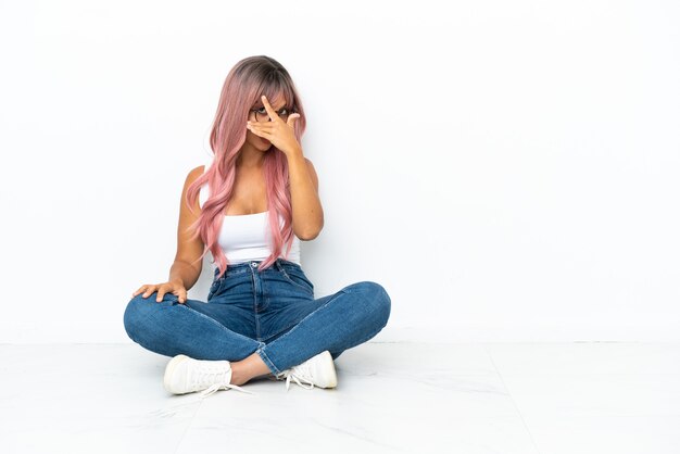 Young mixed race woman with pink hair sitting on the floor isolated on white background covering eyes by hands and smiling