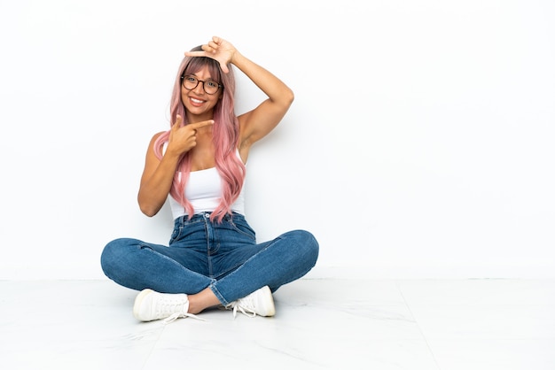 Young mixed race woman with pink hair sitting on the floor isolated on white background focusing face. Framing symbol