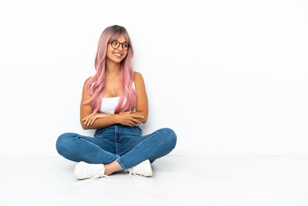 Молодая женщина смешанной расы с розовыми волосами сидит на полу на белом фоне, глядя вверх, улыбаясь