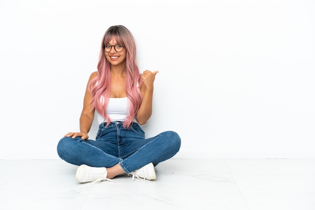 Young mixed race woman with pink hair sitting on the floor isolated on white background pointing to the side to present a product