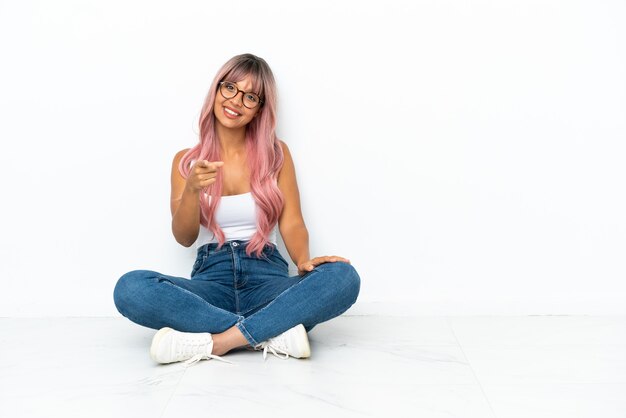 Young mixed race woman with pink hair sitting on the floor isolated on white background points finger at you with a confident expression