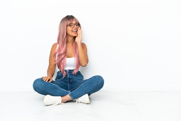 Young mixed race woman with pink hair sitting on the floor isolated on white background shouting with mouth wide open to the lateral