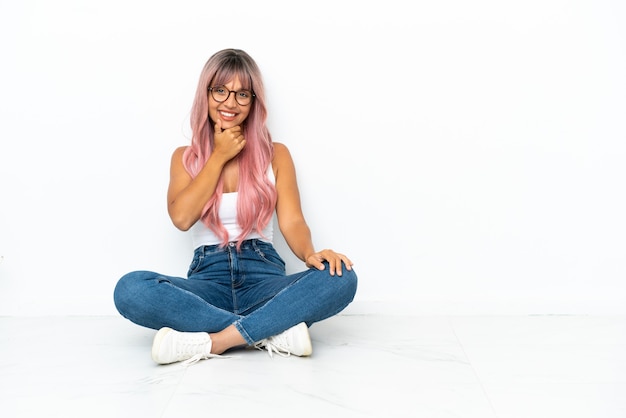 Young mixed race woman with pink hair sitting on the floor isolated on white background with glasses and smiling