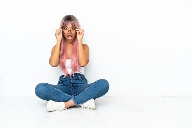 Young mixed race woman with pink hair sitting on the floor isolated on white background with glasses and surprised