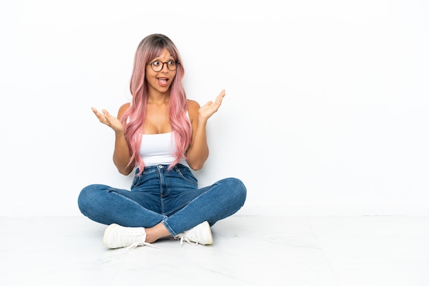 Young mixed race woman with pink hair sitting on the floor isolated on white background with surprise facial expression