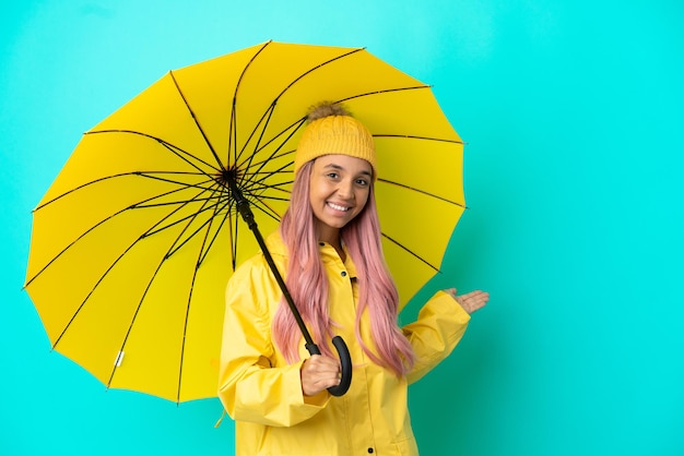Young mixed race woman with rainproof coat and umbrella extending hands to the side for inviting to come