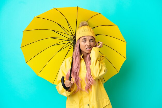 Young mixed race woman with rainproof coat and umbrella frustrated and covering ears