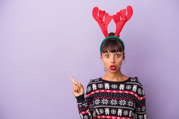 Young mixed race woman with reindeer hat celebrating Christmas isolated on purple background  pointing to the side