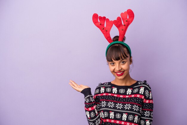 Young mixed race woman with reindeer hat celebrating Christmas isolated on purple background  showing a copy space on a palm and holding another hand on waist.