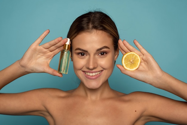 A young model holds a serum with vitamin c and half a lemon in her hands on a blue background