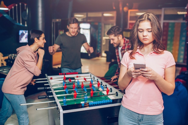 분홍색 셔츠에 젊은 모델 손에 전화를 개최. 행복 한 젊은 팀 재생 방에 테이블 축구 게임. 남자는 여자를 향해 서 있습니다.