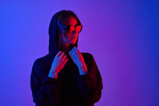 Young model posing in a trendy neon light. Woman in sunglasses and hoodie with colorful light effects. Disco style. Gel filter.