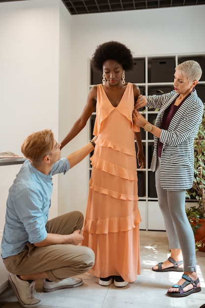 Young model wearing a new orange dress standing next to two fashion designers in a studio