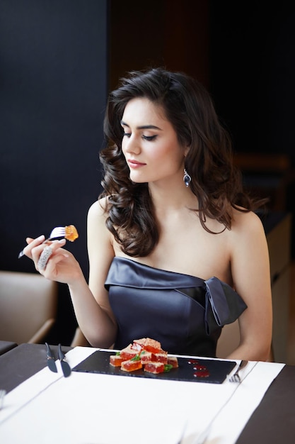 Молодая модель с уложенной прической и макияжем в элегантном платье подает и пробует десерт в ресторане