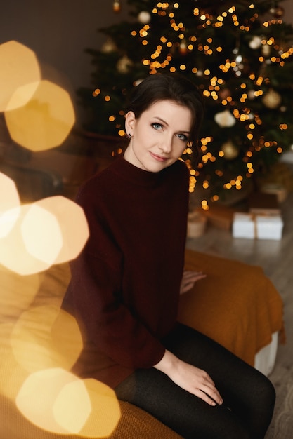 Young model woman with bright makeup wearing red knitted dress posing on the bed with festive lights in the background.