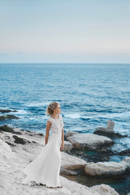 Photo young model woman with perfect body in stylish long white dress posing on the rocks on the seacoast at cyprus