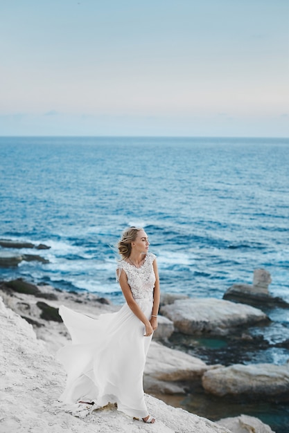 Young model woman with perfect body in stylish long white dress posing on the rocks on the seacoast at Cyprus