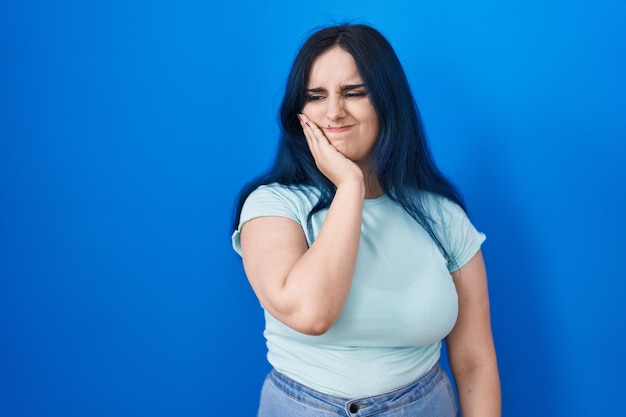 Young modern girl with blue hair standing over blue background touching mouth with hand with painful expression because of toothache or dental illness on teeth dentist