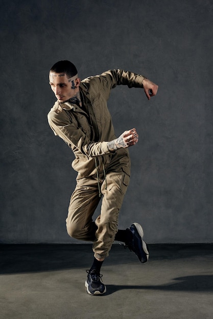 Young modern guy with tattooed body and face, earrings, beard. Dressed in khaki overalls and black sneakers. He is dancing against gray studio background. Dancehall, hip-hop. Full length, copy space