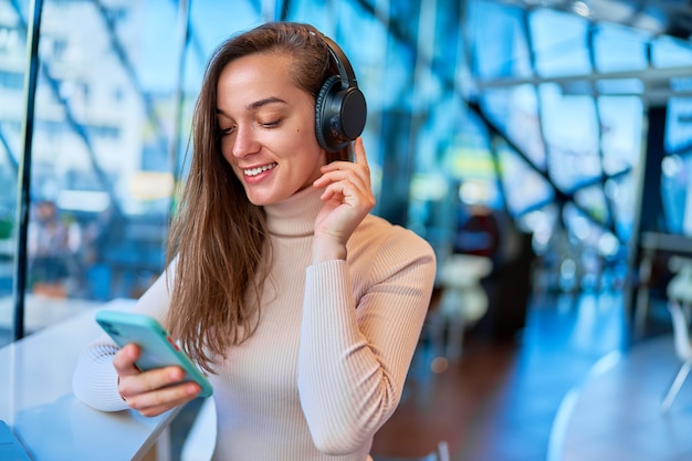 Young modern happy joyful woman wearing wireless headphones using smartphone for watching video online while sitting at cafe