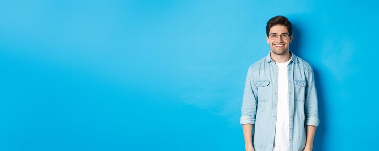 Photo young modern man in glasses and casual outfit standing against blue background smiling happy at came
