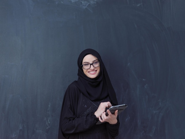 young modern muslim business woman using tablet computer wearing glasses and hijab clothes in front of black chalkboard