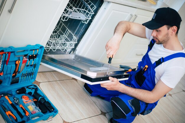 Young modern serviceman in worker suit during the repairing of\
the dishwasher on the domestic kitchen.