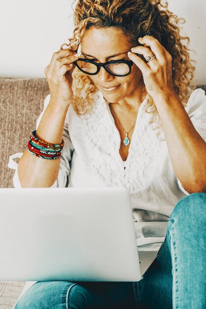 Foto donna giovane e moderna a casa che lavora felice su un laptop seduta sul divano persona che lavora online che usa il computer con un sorriso belle donne che navigano sul web da sole a casa con gli occhiali e divertimento