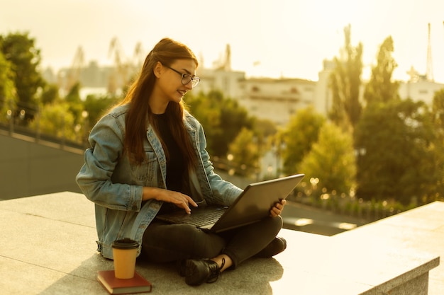 Young modern woman student in a denim jacket sitting and uses laptop outdoor. Distance learning. Modern youth concept.