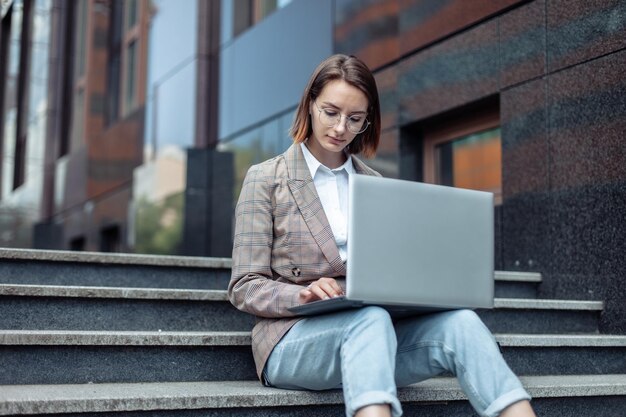 Young modern woman student or freelancer sits on the stairs and uses a laptop Distance learning or work