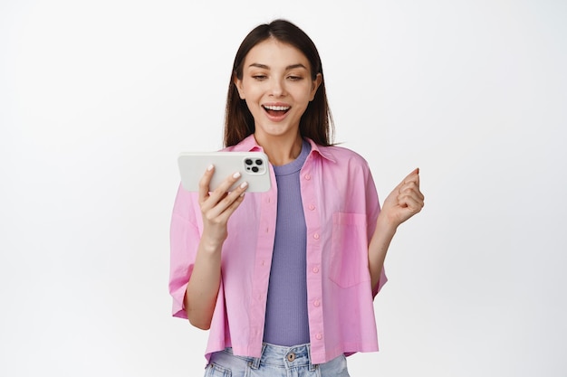 Giovane donna moderna che guarda sul telefono cellulare guardando lo smartphone con uno sfondo bianco di espressione faccia sorridente allegra