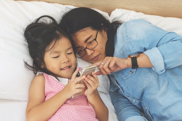 若いお母さんと彼女の小さな娘は家庭のベッドでスマートフォンを使用しています