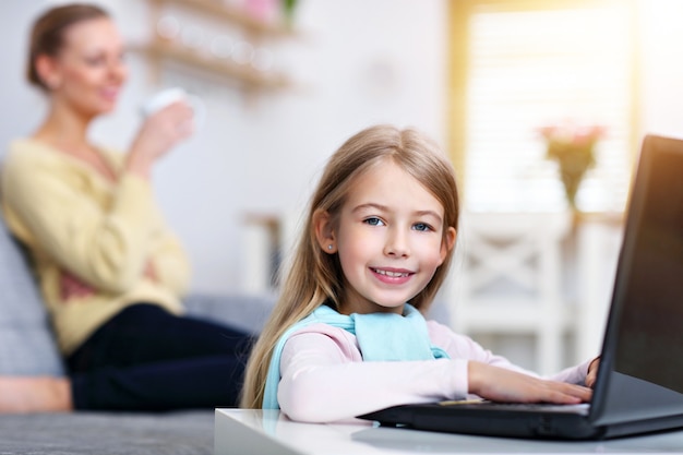 young mom and her little daughter using laptop and sitting on sofa at home