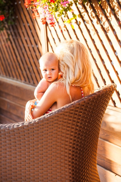 The young mom in a swimsuit sitting in a wicker armchair and hugging baby, view from behind