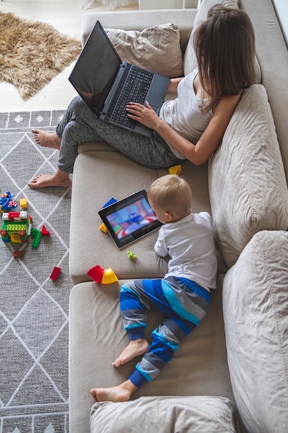 Young mom works at home sitting on couch with laptop while her little kid plays with tablet