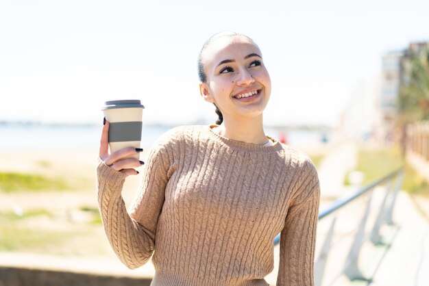 微笑みながら見上げる屋外で持ち帰り用のコーヒーを保持している若いモロッコの女の子